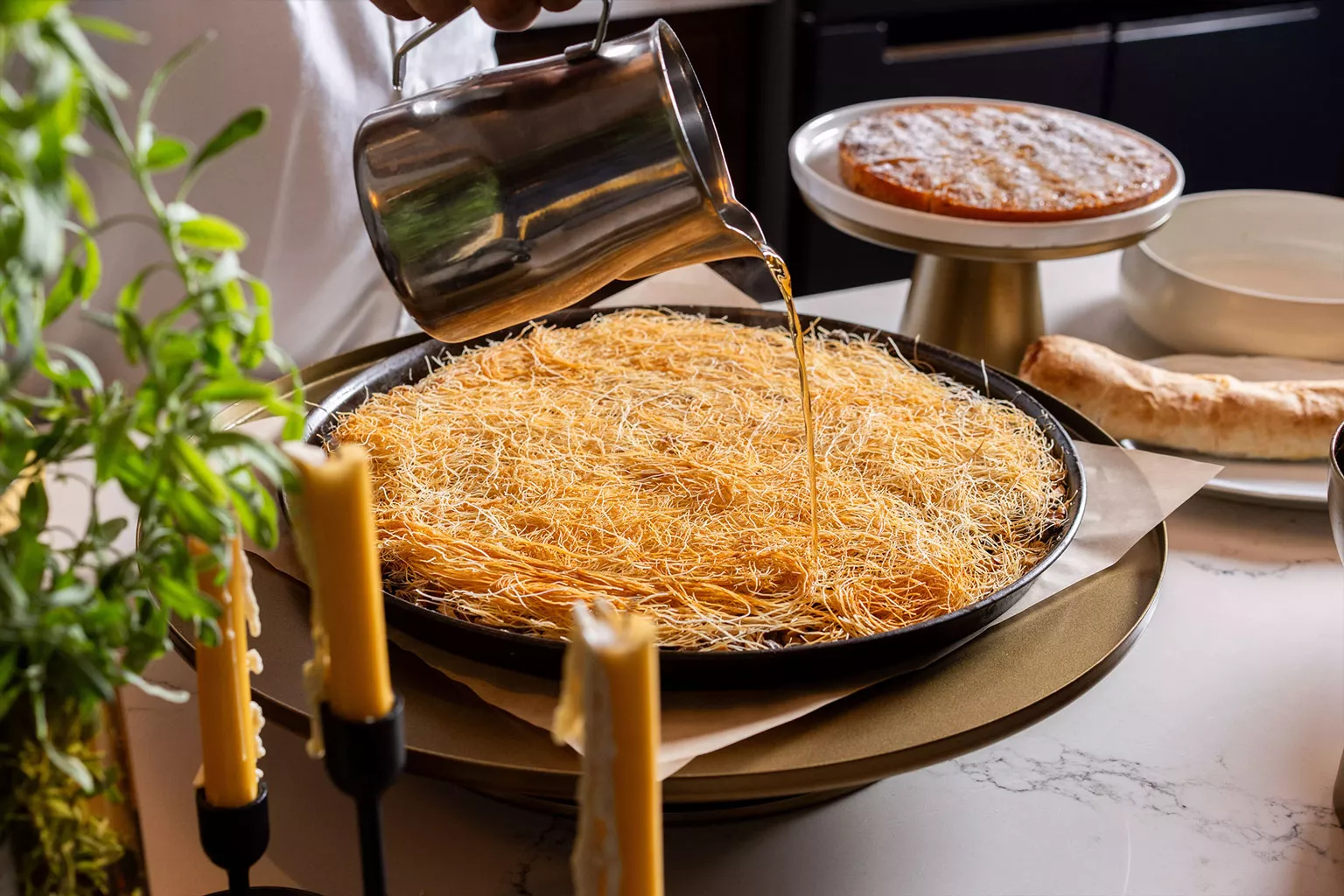 טעימות השף יד מוזגת סירופ חם מקנקן מתכת על כנאפה טרייה במחבת רחבה. צילום תקריב המדגיש את מרקם האטריות, הברק של הסירופ והכנה מוקפדת של קינוח מסורתי.