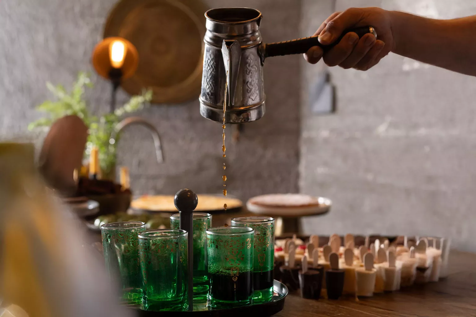 יד מוזגת קפה חם מקנקן מתכת לכוסות זכוכית ירוקות, על שולחן אירוח עם קינוחים אישיים ברקע. צילום תקריב המדגיש תנועה, חום ואווירה אינטימית.