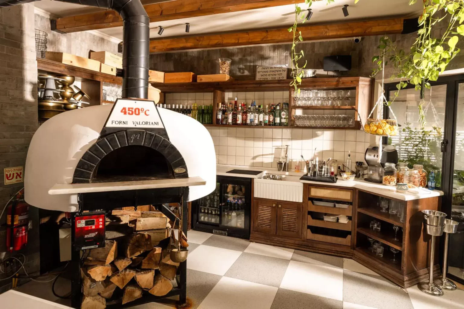 טאבון עצים מקצועי לאפייה בחום גבוה, משולב במטבח פתוח עם משטחי עבודה, מדפי בקבוקים, כלי מטבח וצמחייה. החלל מעוצב בסגנון חמים עם עץ, תאורה טבעית ואווירת עבודה ואירוח.