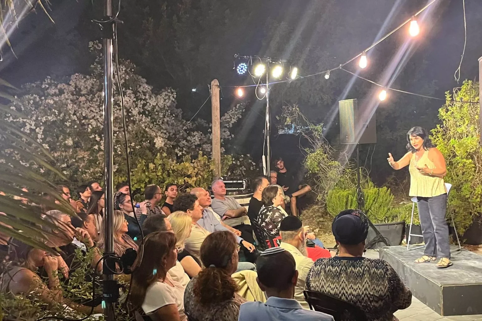 קהל יושב באירוע חוץ בערב וצופה במרצה על במה קטנה עם תאורה