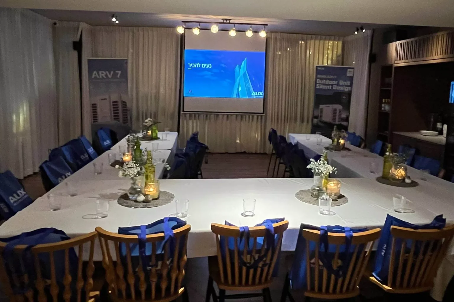 חדר אירועים ערוך לישיבה סביב שולחנות עם מסך מקרן למצגת ותאורה חמה