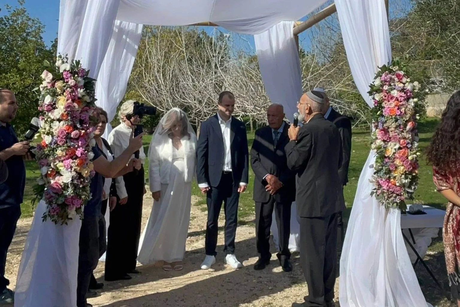 טקס חופה בחתונה בחצר פתוחה עם קישוטי בד לבן ועמודי פרחים