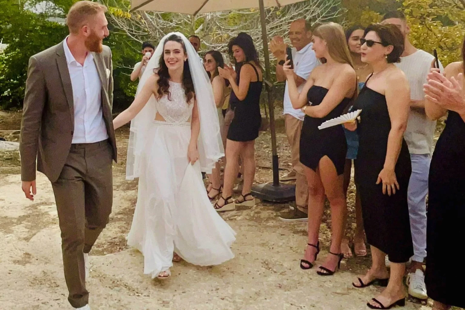 חתן וכלה נכנסים לחתונה בחוץ והאורחים מוחאים כפיים ומצלמים